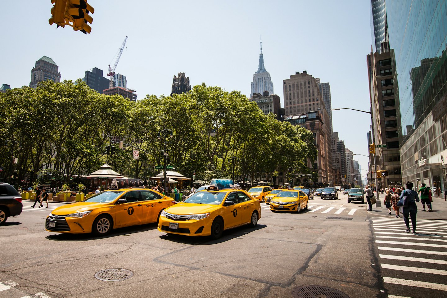 Yellow taxis drive down a busy city street with pedestrians and trees.