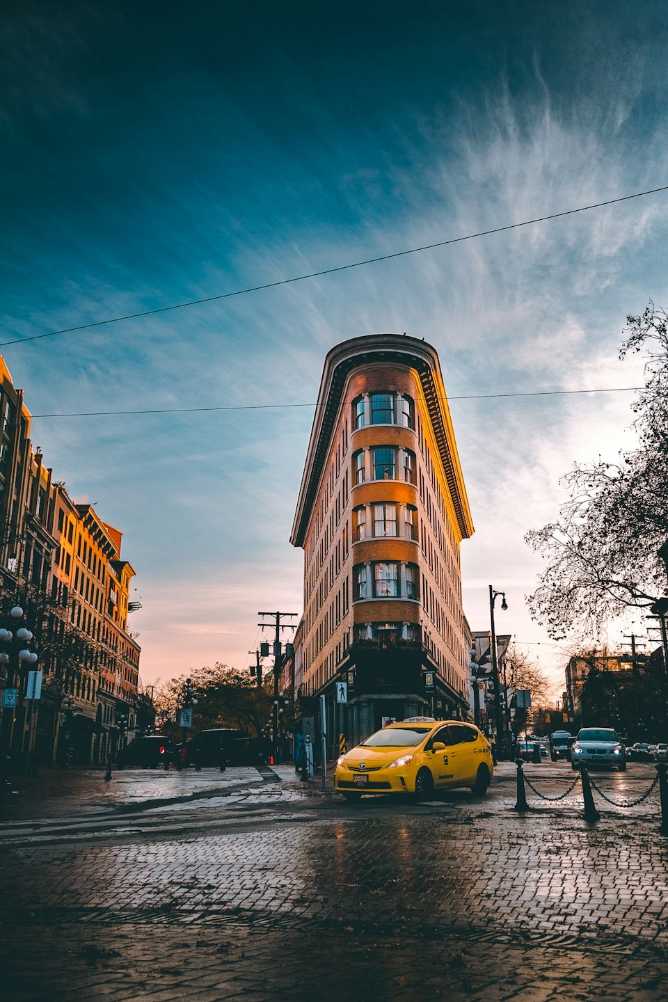 A yellow taxi drives along a wet cobblestone city street.